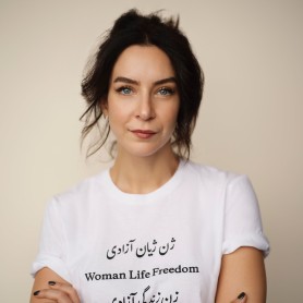 A picture of a woman named Tara Aghdashloo. She is portrayed from the chest up against a plain, light beige background. She has dark, wavy hair loosely pulled back, allowing a few strands to frame her face. She has striking blue eyes and a composed, confident expression with a subtle smile. Her arms are crossed and she is wearing a white t-shirt with rolled sleeves, featuring black text in four languages that states “Woman Life Freedom”.