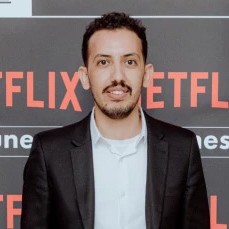 A picture of a man named Mohamed Echkouna. He stands against a dark backdrop with the word “NETFLIX” repeated in bright red letters. He has short dark hair and a trimmed beard, and he is dressed in a sharp, black suit with a white dress shirt beneath it. He is smiling gently and looking directly into the camera.