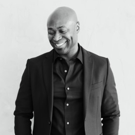 A black-and-white picture of a man named Anslem Richardson against a plain light-colored wall. He is smiling warmly while looking downwards. He is dressed in a dark, tailored suit with a matching button-up shirt underneath. The suit jacket has a simple, sleek design with a notched lapel and the shirt is neatly buttoned up to the collar. The man is standing with his hands casually resting in his pockets. His head is bald, and his facial expression exudes a serene and content demeanor.