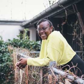A picture of a woman named Jennifer Drake. She is shown leaning against a metallic fence covered with dry vines, smiling warmly at the camera. She sports short, braided hair and is wearing glasses and a vibrant yellow top with long sleeves.
