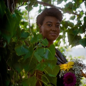 A picture of a woman named Natalie Harris. Natalie is standing amidst dense green foliage with sunlight filtering through the leaves. She has short curly hair and wears a pair of simple hoop earrings. Natalie is holding a vibrant bouquet of flowers, including yellow and purple blooms, along with white baby's breath. Her expression is calm and gentle, with a slight smile on her lips.
