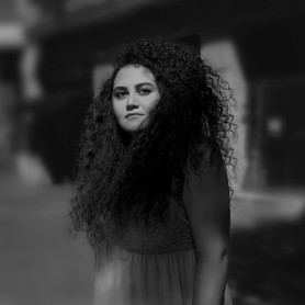 A black-and-white picture of a woman named Ashtar Alahmad. She is standing outdoors with an indistinct, blurred background. She has long, voluminous, curly dark hair that frames her face down to her shoulders. Ashtar is wearing a sleeveless top or dress featuring intricate patterns across the chest area. She gazes directly at the camera with a calm, composed expression.