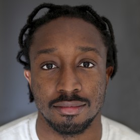 A picture of a man named Rhys Aaron Lewis. He has short, twisted hair, a short beard, and a mustache. He is wearing a light gray T-shirt. The man's face fills most of the frame, capturing a neutral expression with a slight hint of a smile. His eyes are looking directly into the camera, and the background is a smooth gradient of dark gray.