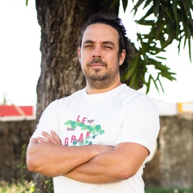A picture of a man named Nicolas Sery. The man is standing outdoors in front of a large tree trunk with green leaves partially visible above his head. He has fair skin, dark hair pulled back, and a subtle facial hair stubble. He is wearing a white t-shirt that has colorful text and graphical elements on it. He has his arms folded across his chest and is looking at the camera with a confident, slight smile.