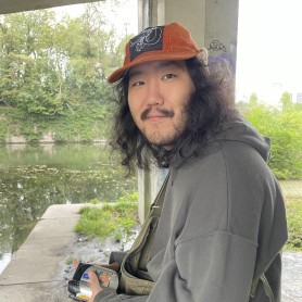 A picture of a man named Kyhynngy Oyuur. He is outdoors wearing a rust-colored hat, sitting under a concrete structure by a calm body of water. He has long curly hair and a mustache, and he is wearing a dark gray hoodie and olive-green overalls. He is holding a camcorder in his hand and is looking directly at the camera with a gentle smile.
