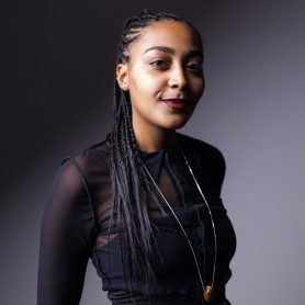 A picture of a woman named Alicia Mendy. She stands confidently against a gradient gray background with a bold red lip. She has long, braided hair that is pulled back neatly. She is wearing a black, long-sleeved, sheer top that reveals another layer underneath paired with a long, thin necklace with a pendant hanging around her neck. She has a slight, poised smile, and her gaze is directed towards the camera.