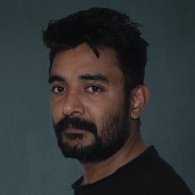 A picture of a man named Niranjan Raj Bhetwal. He is against a plain, dark gray background facing slightly to his left, with his head turned towards the camera. He has short, dark hair that is styled messily. His facial hair consists of a thick, full beard and mustache. He is wearing a black t-shirt and has a calm, serious expression on his face.
