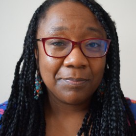 A picture of a woman named Hachimiya Ahamada. The image is a close-up portrait of the woman facing the camera. She has long black braids that frame her face and extend beyond the visible part of the image. She wears red-framed glasses and a gentle serious expression with a slight smile, lending a composed and calm demeanor.