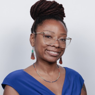 A headshot of Zendra. She is a Black woman, her hair is locked and tied into a bun. She is wearing glasses, earrings, a necklace, and a blue dress.