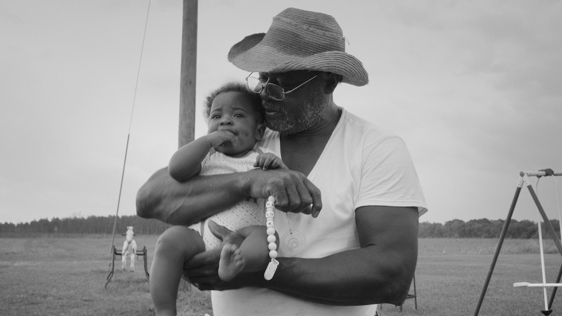 A black-and-white photo of a Black man with a straw-brimmed hat holding a baby in his arms. The baby is looking off into the distance with a concerned expression. The man is looking at the baby with care in his eyes.