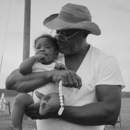 A black-and-white photo of a Black man with a straw-brimmed hat holding a baby in his arms. The baby is looking off into the distance with a concerned expression. The man is looking at the baby with care in his eyes.