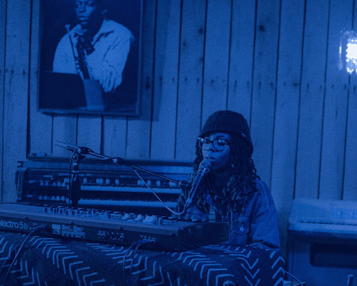 A blue-colorized image of Kayla Childs at Turtle Studios. She is in front of a keyboard, her eyes are closed and she is singing into a microphone.