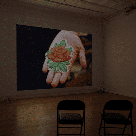 A photo of an empty gallery space where a film is showing. In the film an outstretched hand is showing a sewn object, which appears to be a flower.