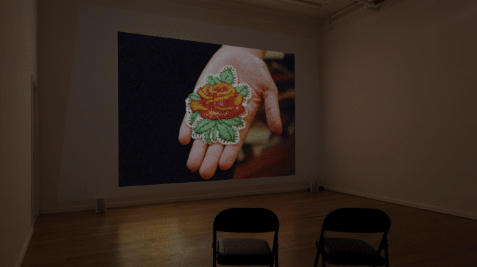 A photo of an empty gallery space where a film is showing. In the film an outstretched hand is showing a sewn object, which appears to be a flower.