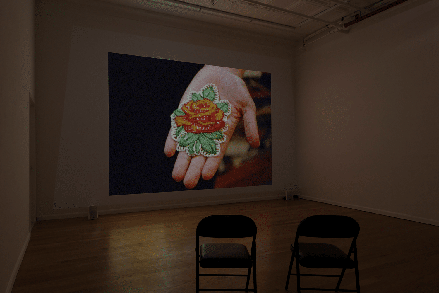 A photo of an empty gallery space where a film is showing. In the film an outstretched hand is showing a sewn object, which appears to be a flower.