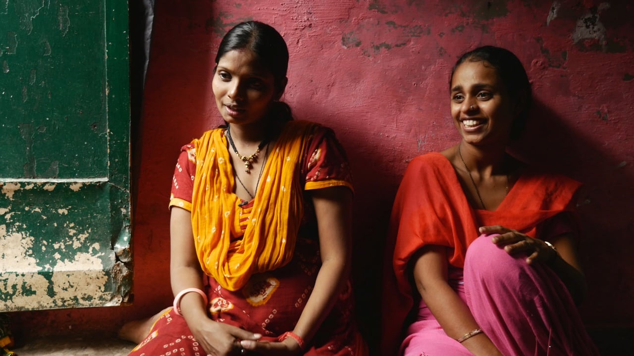 A still from Cosmic Egg shows two Indian women sitting in front of a red painted wall. They are dressed in traditional clothing and are smiling.