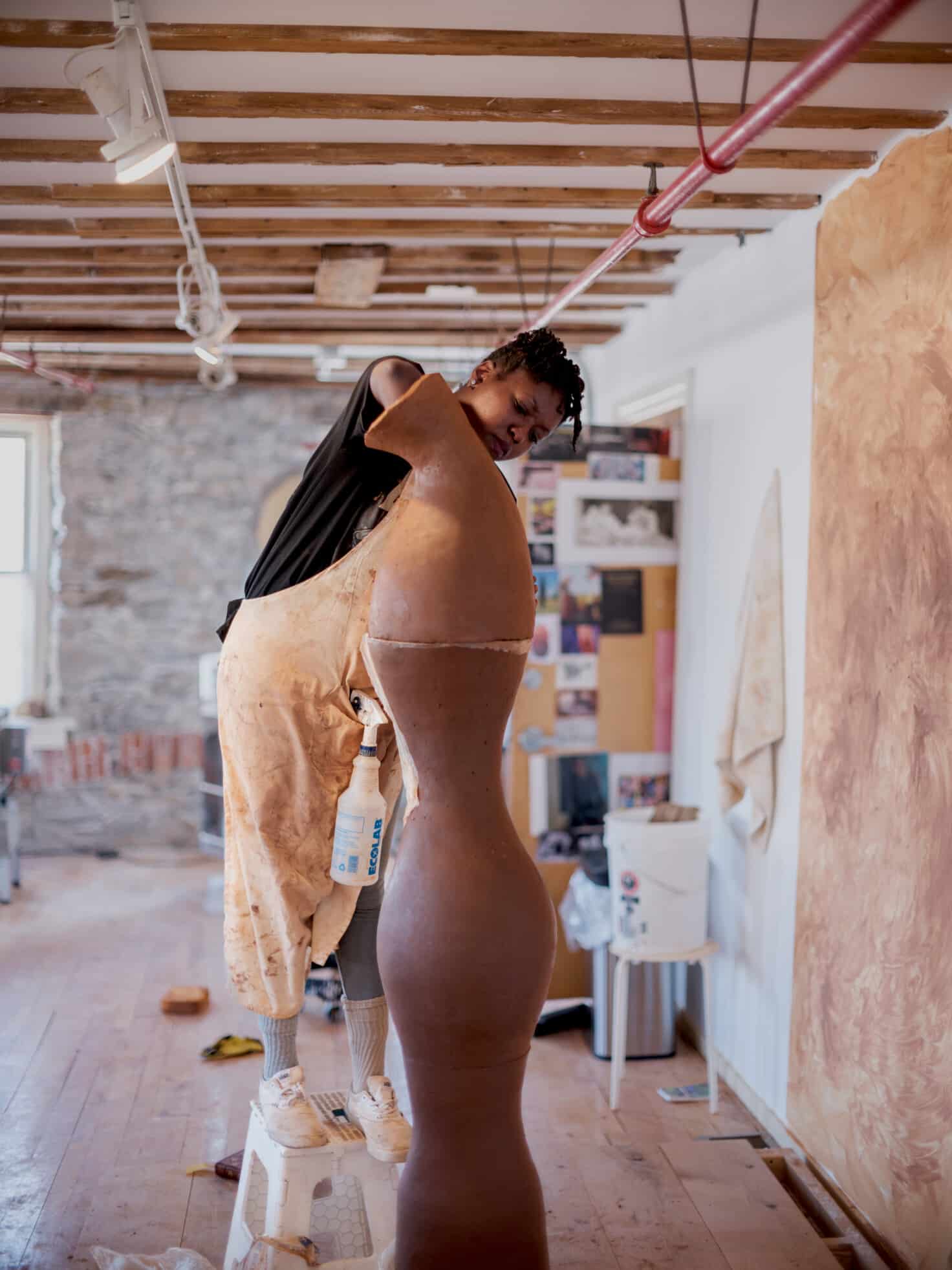 While standing on a stool, Adebunmi reaches into the top structure of a tall sculpture in progress.