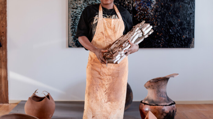 Adebunmi Gbadebo stares into the camera while holding pieces of wood. She is standing up straight, surrounded by many different vases she's created.