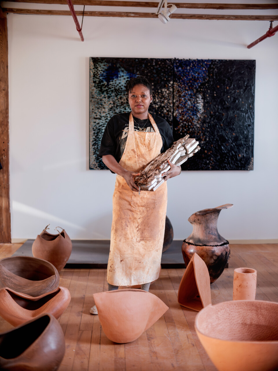 Adebunmi Gbadebo stares into the camera while holding pieces of wood. She is standing up straight, surrounded by many different vases she's created.