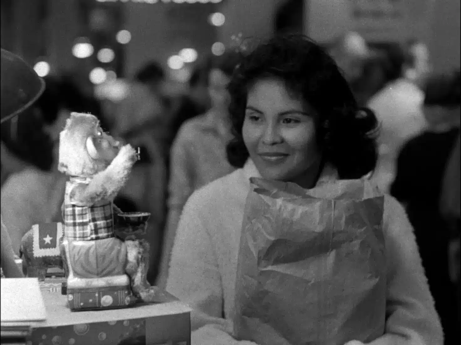 In a black and white still from “The Exiles,” a woman holding a paper bag smiles at a drumming monkey statue.