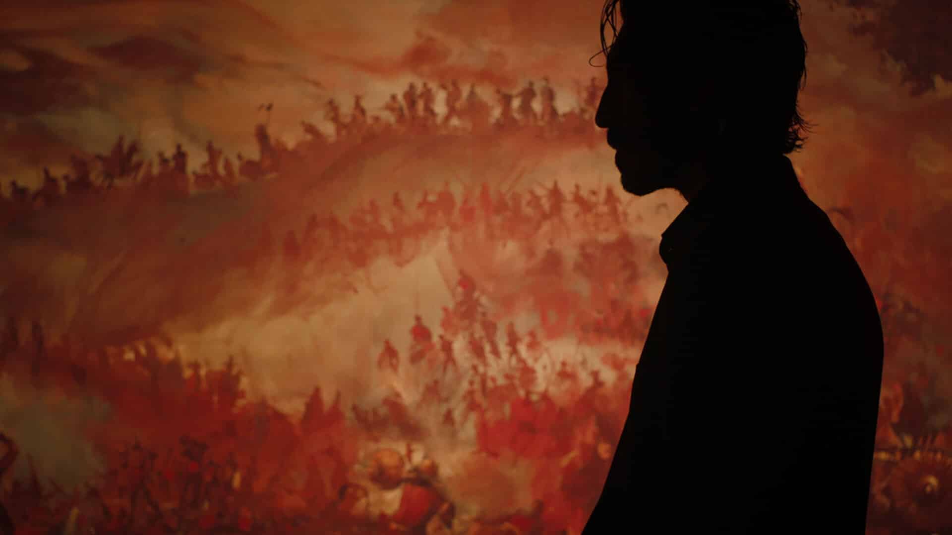 A still photo from “Monkey Man” (2024) showing an opaque side profile of Kid (played by Dev Patel) against a background of wallpaper depicting war.