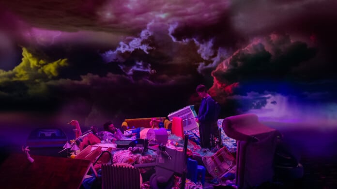 A still image from “Problemista” (2023) featuring a young man stares at a pile of his belongings. Above him is a sky with stormy clouds.