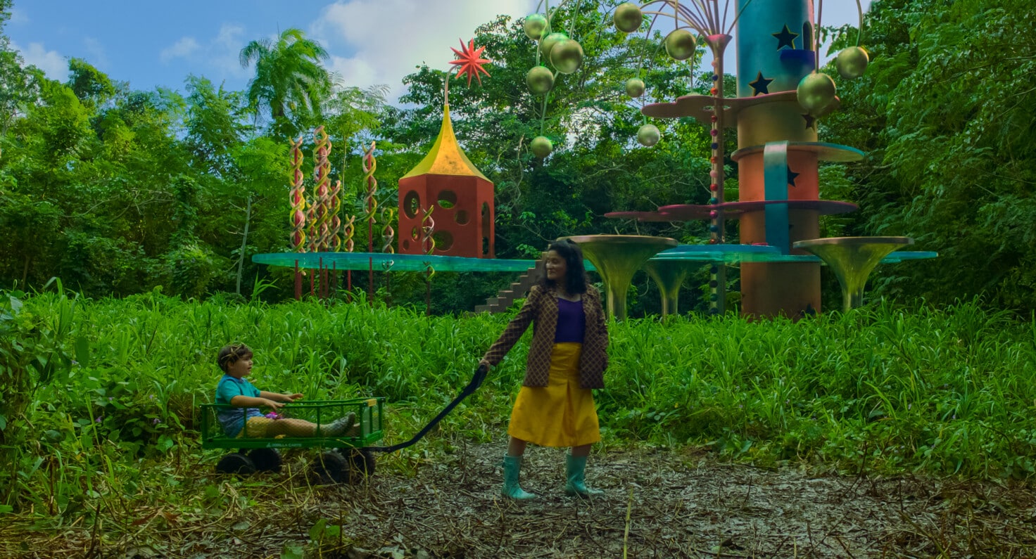 A still image from “Problemista” (2023) showing a young boy in a wagon being pulled by a young woman in a yellow skirt and green rain boots through a grassy area with a kids playground in the background.