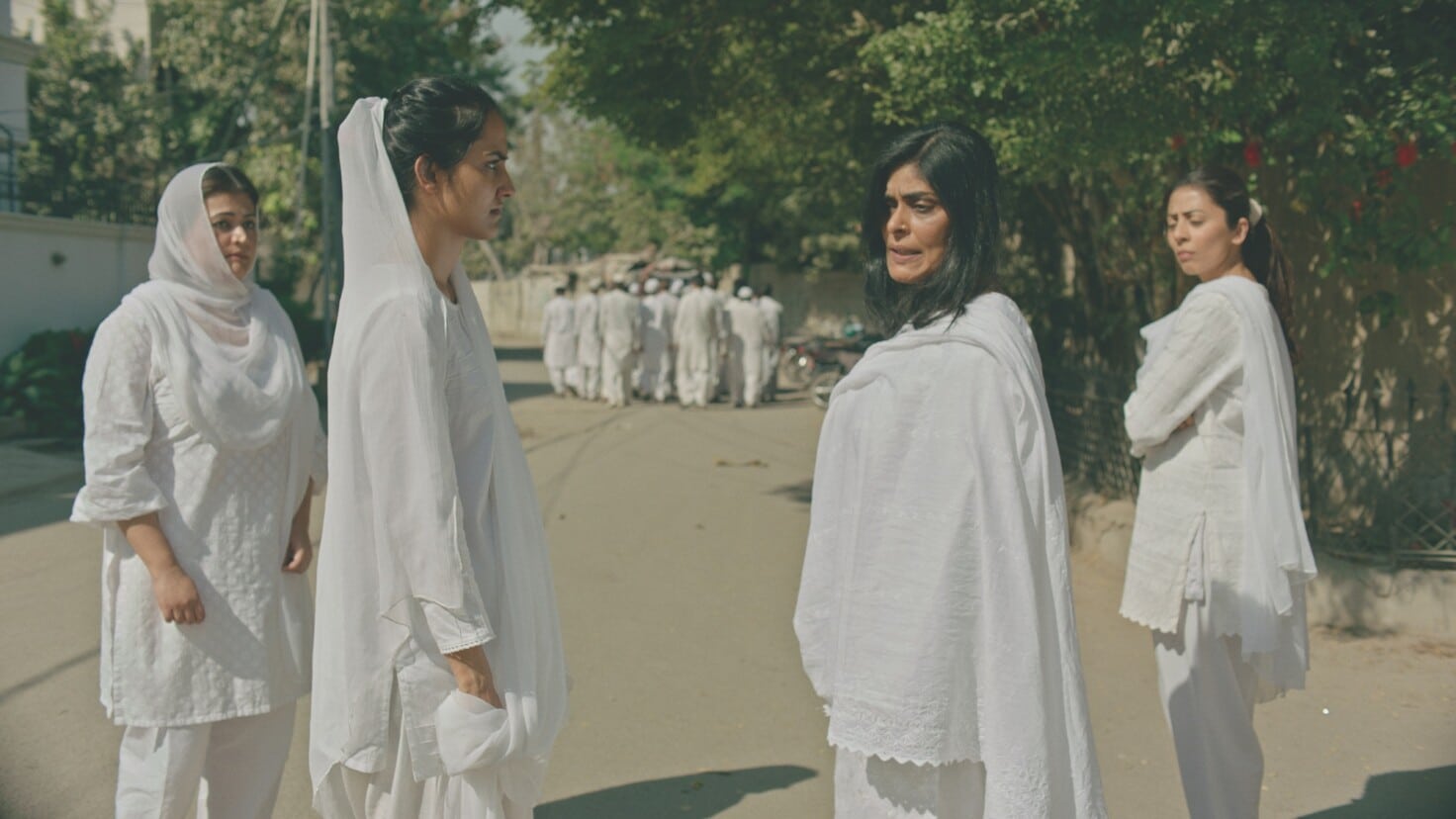 A still image from “The Queen of My Dreams” (2023) showing a young Indian woman in a white saree and head scarf looking at an older Indian woman also wearing a white saree and head scarf. In the middle of the image, another older Indian woman looks at the two women. In the background, a group of men wearing white gowns walk into the distance.