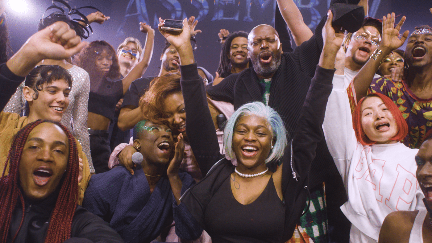 A still from Assembly shows a group of Black and Brown people, they are smiling and laughing, their arms are raised in celebration.