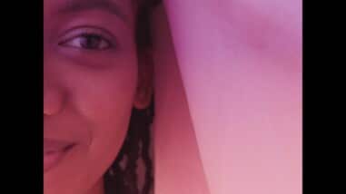A film still from 16 ½ shows half the face of a young Black girl. Her gaze is straight ahead with a gentle smile. A cloth is presumably draped over her head. There is a pink overlay over the entire still.