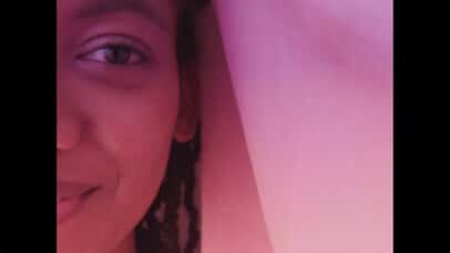 Half of a smiling young black woman's face frame-left looking directly into camera sitting under a bedsheet bathed in pink and purple lights.