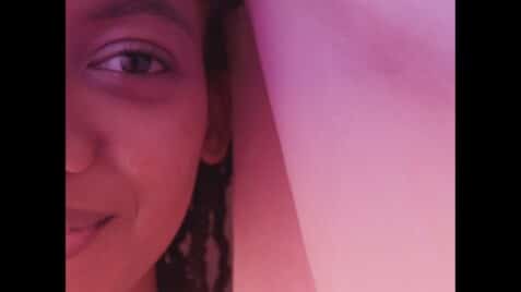 A film still from 16 ½ shows half the face of a young Black girl. Her gaze is straight ahead with a gentle smile. A cloth is presumably draped over her head. There is a pink overlay over the entire still.