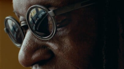 A close-up shot of an elder man wearing round, dark-rimmed glasses. His eyes are mostly closed, and the reflection in his glasses shows a window and a glimpse of the outside world. The lighting is soft, casting gentle shadows on his face, creating an intimate and contemplative tone.