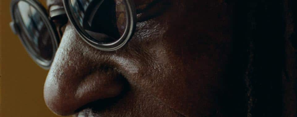 A close-up shot of an elder man wearing round, dark-rimmed glasses. His eyes are mostly closed, and the reflection in his glasses shows a window and a glimpse of the outside world. The lighting is soft, casting gentle shadows on his face, creating an intimate and contemplative tone.