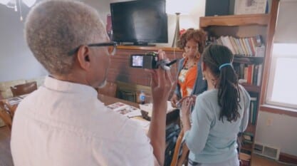 A film still from A New Voice shows two Black women in conversation presumably in a dining room with wooden furniture. The older woman is wearing an orange floral dress and gold and orange pendant earrings. The other woman with her back to the camera has locs tied together with a blue hair band. There is an older Black man recording the women with a camcorder.