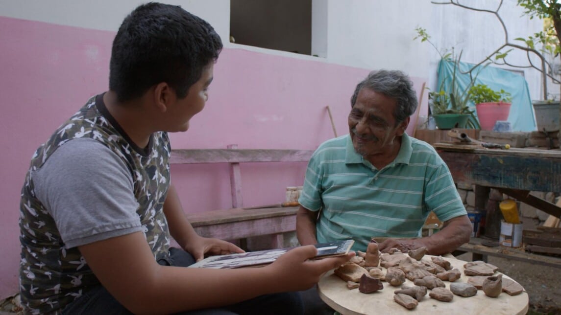 A film still from Binnigula’sa’ (Ancient Zapotec People) shows a young boy in conversation with an older man. The boy is looking at the man who looks at a photo the boy holds in his hands. They are seated outside around a table with small crafted figures.