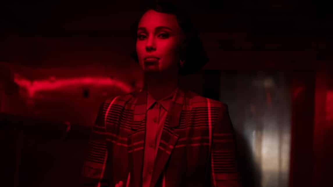 A film still from BLKNWS: Terms & Conditions shows a woman standing in a room illuminated by red lights. There is a thick dark ink that goes from the bottom of her lip to her chin. Her hands are stacked on top of each other palm up in front of her. She is wearing a plaid suit.