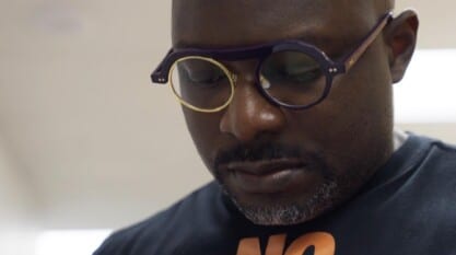 A film still from Bloodlines, Mississippi shows a Black man looking down at something off screen. He is wearing a black t-shirt with an orange graphic and purple circle lensed glasses, the left lens is gold.