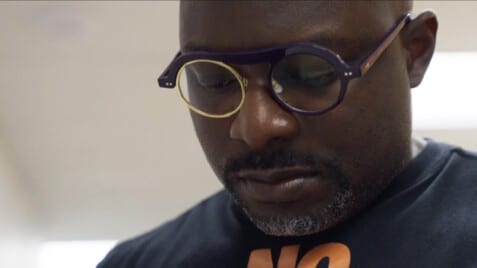 A film still from Bloodlines, Mississippi shows a Black man looking down at something off screen. He is wearing a black t-shirt with an orange graphic and purple circle lensed glasses, the left lens is gold.