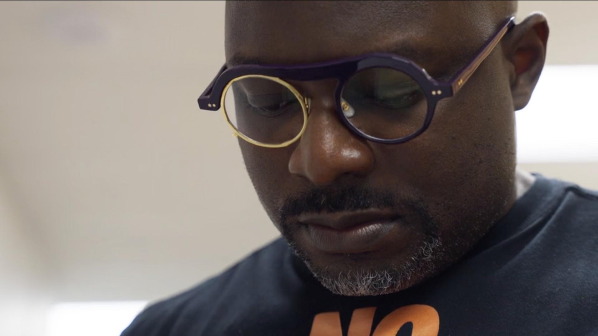 A film still from Bloodlines, Mississippi shows a Black man looking down at something off screen. He is wearing a black t-shirt with an orange graphic and purple circle lensed glasses, the left lens is gold.