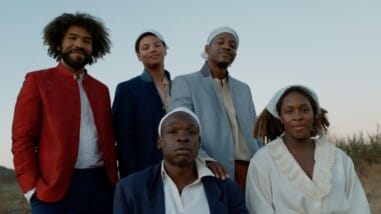 A film still from Boil That Cabbage Down shows a group of Black people posed together in a field with a clear light blue sky. At the center is a man wearing a blue blazer over a white collared shirt. Next to him is a woman wearing a white shirt with a frilly collar. Behind them stand three Black men also in blazers. The man on the left stands out, wearing a red blazer and an afro. He is the only person without a white scarf tied on their head.