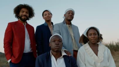 six individuals stand and sit together in a serene, open landscape under a pale blue sky. The group, dressed in 18th-century-inspired attire, exudes quiet strength and unity. Earth-toned and pastel garments, including head wraps and buttoned jackets, reflect a historical aesthetic rooted in resilience and grace. The central seated figure gazes solemnly ahead, flanked by others whose expressions range from serene to introspective. The soft, natural lighting and dry brush backdrop suggest early morning or dusk, evoking themes of endurance, kinship, and heritage.