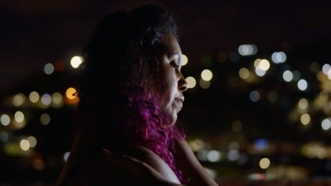 A film still from Brick by Brick shows a woman with a soft gaze looking into the distance at night, her hand resting gently on her opposite shoulder in a self-embrace. She has curly hair that fades from black to hot pink. The background is blurred, filled with soft dots of light.
