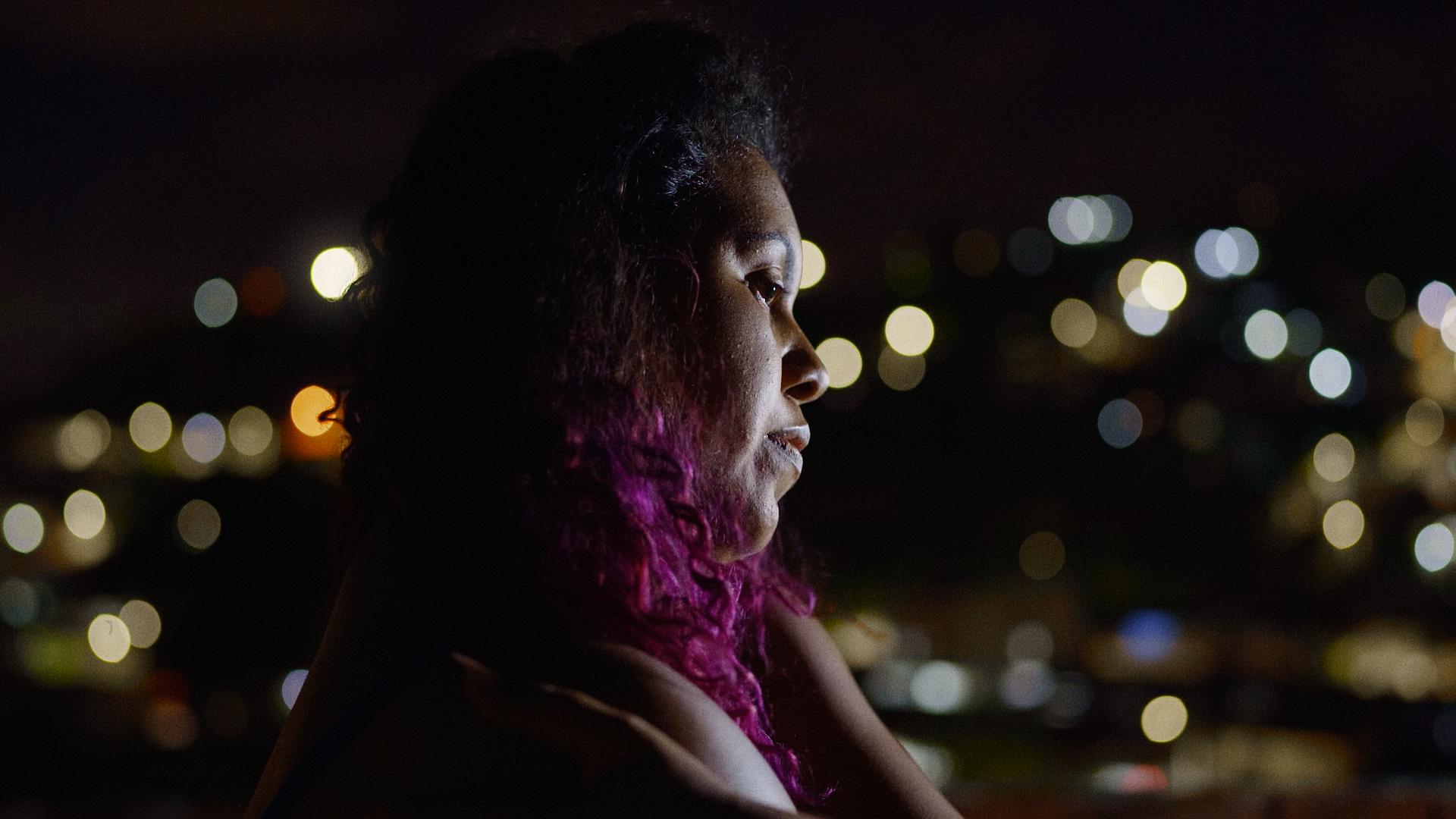 A film still from Brick by Brick shows a woman with a soft gaze looking into the distance at night, her hand resting gently on her opposite shoulder in a self-embrace. She has curly hair that fades from black to hot pink. The background is blurred, filled with soft dots of light.