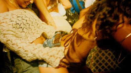 A film still from Bubbling Baby shows two women in a crowd. The woman on the left is wearing a white knitted off the shoulder top. She has her hands on the waist of the other woman who is bent over in front of her. The other woman is wearing a silk tank top tied in the back and jean shorts.