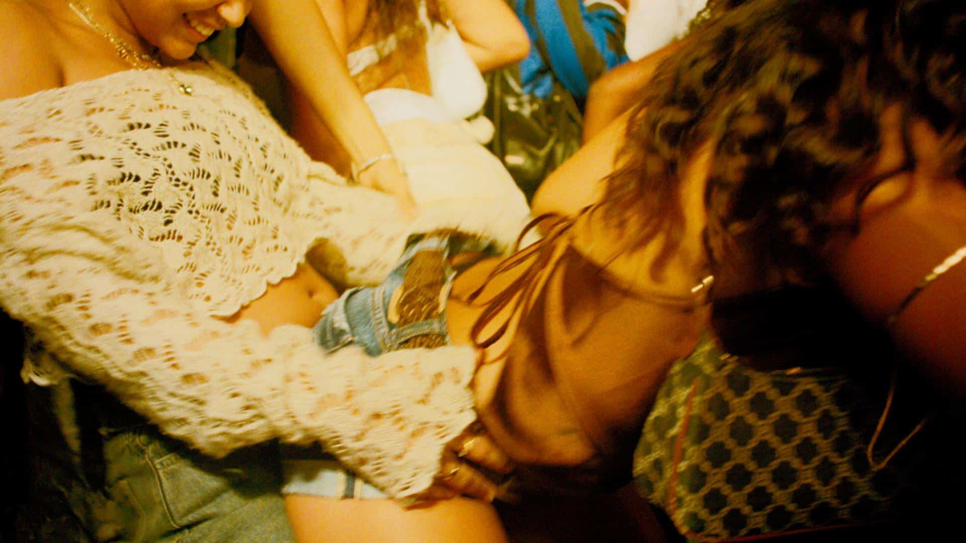 A film still from Bubbling Baby shows two women in a crowd. The woman on the left is wearing a white knitted off the shoulder top. She has her hands on the waist of the other woman who is bent over in front of her. The other woman is wearing a silk tank top tied in the back and jean shorts.