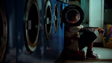 A film still from Budget Paradise shows a series of blue washing machines in a laundromat. The second to last washing machine door is open. The glass on the door blurs the face of a young person sitting on a stool with their back against the washer looking out into the distance.