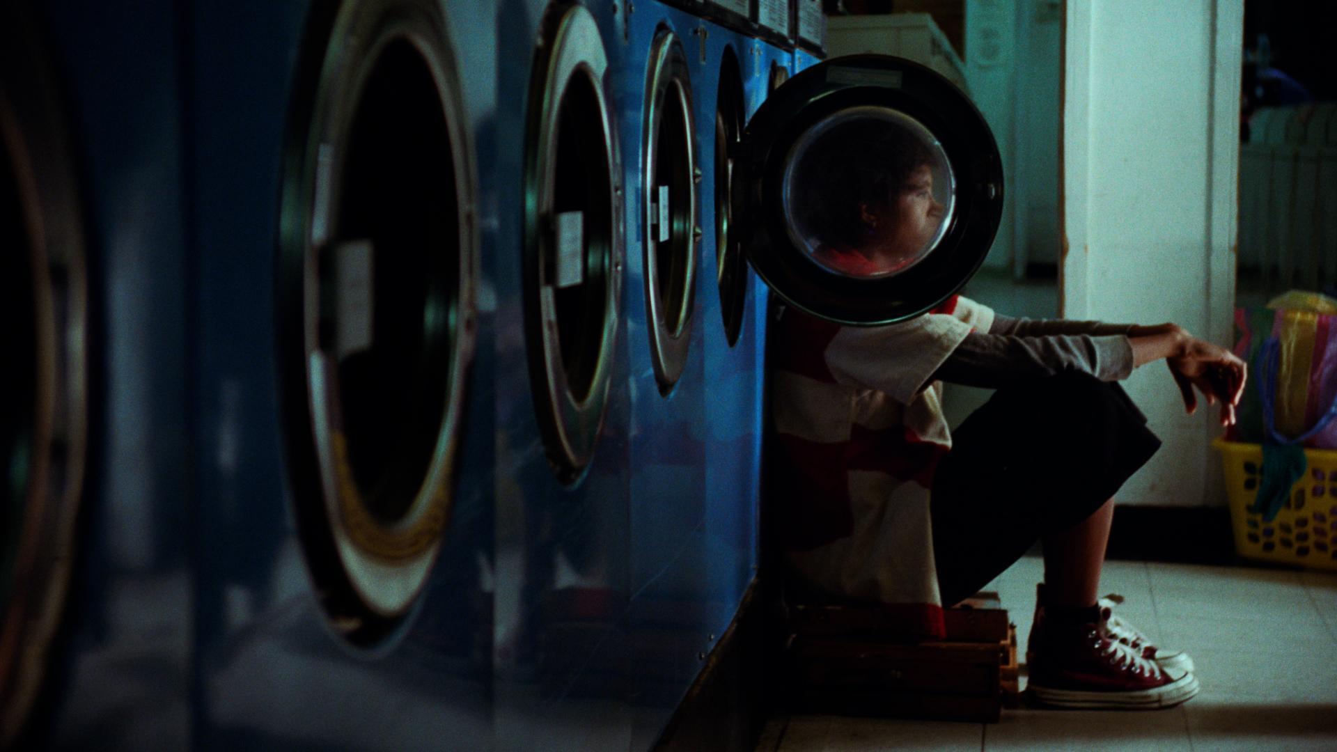 A film still from Budget Paradise shows a series of blue washing machines in a laundromat. The second to last washing machine door is open. The glass on the door blurs the face of a young person sitting on a stool with their back against the washer looking out into the distance.