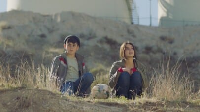 A girl and a boy sit on a grassy hill, a makeshift soccer ball between them. They face the distance, framed by rocky terrain and industrial tanks in the background, evoking a quiet, reflective mood.