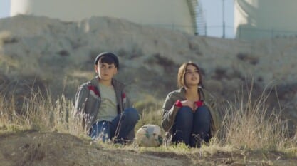 A film still from Bukra shows a young Syrian boy and girl sitting in a field. Both of their gazes in the distance. Between them is a makeshift soccer ball made with different fabrics, held together with bands.